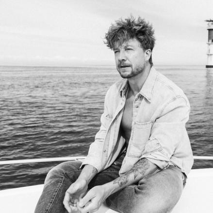 Samu Haber auf einem Boot, schwarz-weiß fotografiert. Im Hintergrund ist ein Leuchtturm zu sehen, sein Hemd ist offen, und er blickt entspannt in die Ferne.