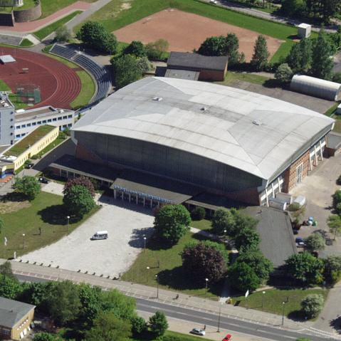 Luftaufnahme der Sport- und Kongresshalle mit großem, geschwungenem Dach. Vor dem Eingang befindet sich ein großer, gepflasterter Vorplatz. Im Hintergrund sind Sportplätze, Grünflächen und umliegende Gebäude zu sehen.