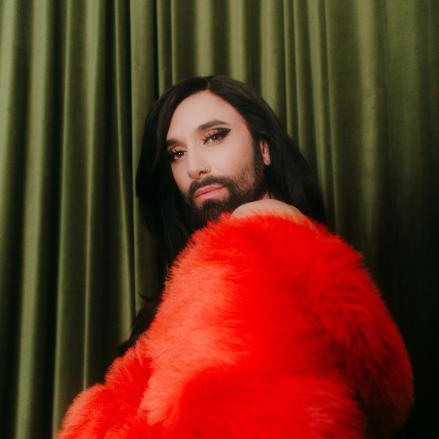 Conchita Wurst in einem auffälligen roten Federmantel. Die Künstlerin blickt ausdrucksstark in die Kamera, das dramatische Styling und Licht betonen die Szene. Foto: Hanna Fasching.