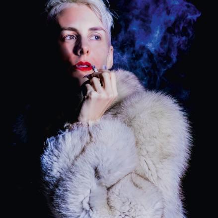 Das Bild zeigt Lisa Eckhart in einem dramatischen, halbdunklen Setting. Sie trägt einen weißen Pelz und hält eine Zigarette, während sie mit einem nachdenklichen Ausdruck leicht zur Seite blickt. Ihr Gesicht ist nur halb beleuchtet.