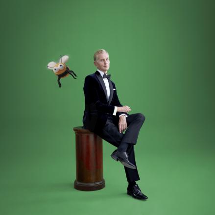 Das Bild zeigt Max Raabe auf einem Hocker sitzend vor einem grünen Hintergrund. Er trägt seinen typischen, eleganten Anzug und blickt in die Kamera, dabei strahlt er eine ruhige, charismatische Präsenz aus. Ein Comic-Biene fliegt in der Nähe von Max Raabe.