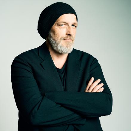 Torsten Sträter steht mit verschränkten Armen und ernst-humorvollem Blick vor dunklem Hintergrund. Er trägt eine schwarze Mütze und dunkle Kleidung, was seinen charakteristischen Stil betont. Sein Blick ist direkt in die Kamera gerichtet und vermittelt eine Mischung aus Selbstbewusstsein und verschmitzter Gelassenheit.