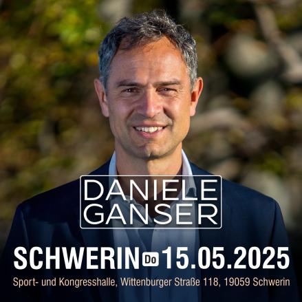 Ein Mann mittleren Alters vor einem verschwommenen natürlichen Hintergrund schaut lächeln in die Kamera. Er trägt ein hellgraues Hemd und ein blaues Sacko.