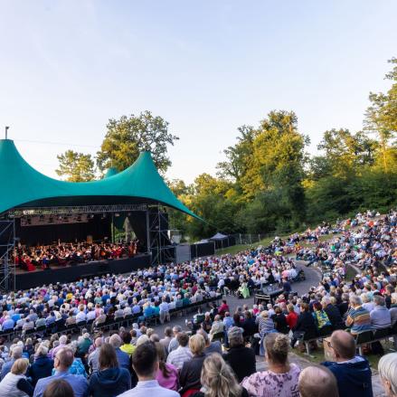 Ein Open-Air-Konzert in einem Amphitheater mit einer großen Zuschauerzahl. Die Bühne hat ein grünes, geschwungenes Dach und ein Orchester spielt vor einem dicht besetzten Publikum. Die Veranstaltung findet bei schönem Wetter inmitten einer grünen, bewaldeten Umgebung statt.