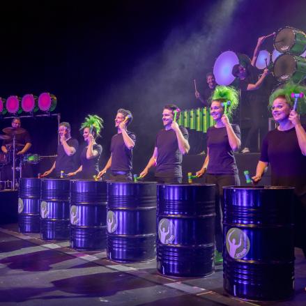 Das Bild zeigt eine Szene von „Greenbeats“, bei der Trommler hinter großen Tonnen stehen, die als Instrumente genutzt werden. Die Tonnen sind in das Bühnenbild integriert und werden durch das lila Licht in Szene gesetzt. 
