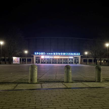 Die Sport- und Kongresshalle bei Nacht mit beleuchteter Beschriftung über dem Eingang. Der Vorplatz ist weitläufig und mit Betonpollern abgegrenzt. Straßenlaternen beleuchten die Szene.