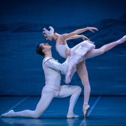 Ein Ballettauftritt von „Schwanensee“ mit zwei eleganten Tänzern. Einer der Tänzer führt eine anmutige Arabeske aus, während der andere in einer dramatischen Pose steht.