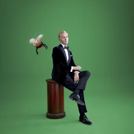 Das Bild zeigt Max Raabe auf einem Hocker sitzend vor einem grünen Hintergrund. Er trägt seinen typischen, eleganten Anzug und blickt in die Kamera, dabei strahlt er eine ruhige, charismatische Präsenz aus. Ein Comic-Biene fliegt in der Nähe von Max Raabe.