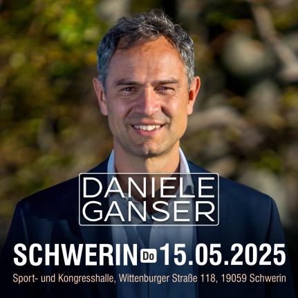 Ein Mann mittleren Alters vor einem verschwommenen natürlichen Hintergrund schaut lächeln in die Kamera. Er trägt ein hellgraues Hemd und ein blaues Sacko.