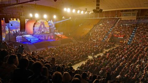 Ein Konzert in einer großen Veranstaltungshalle. Die Bühne ist mit buntem Licht beleuchtet, das Publikum sitzt auf den Rängen und im Innenraum.